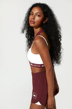A woman modeling a matching athletic set consisting of a white sports bra with a burgundy band and burgundy shorts with a white inner lining, against a plain light gray background.