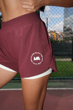 A close-up of a person wearing maroon athletic shorts with a white inner layer and a white circular logo on the left leg, standing on an outdoor court with a chain-link fence in the background.