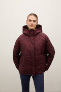 A woman modeling a quilted hooded puffer jacket in a deep burgundy color, paired with navy blue leggings, against a plain off-white background.