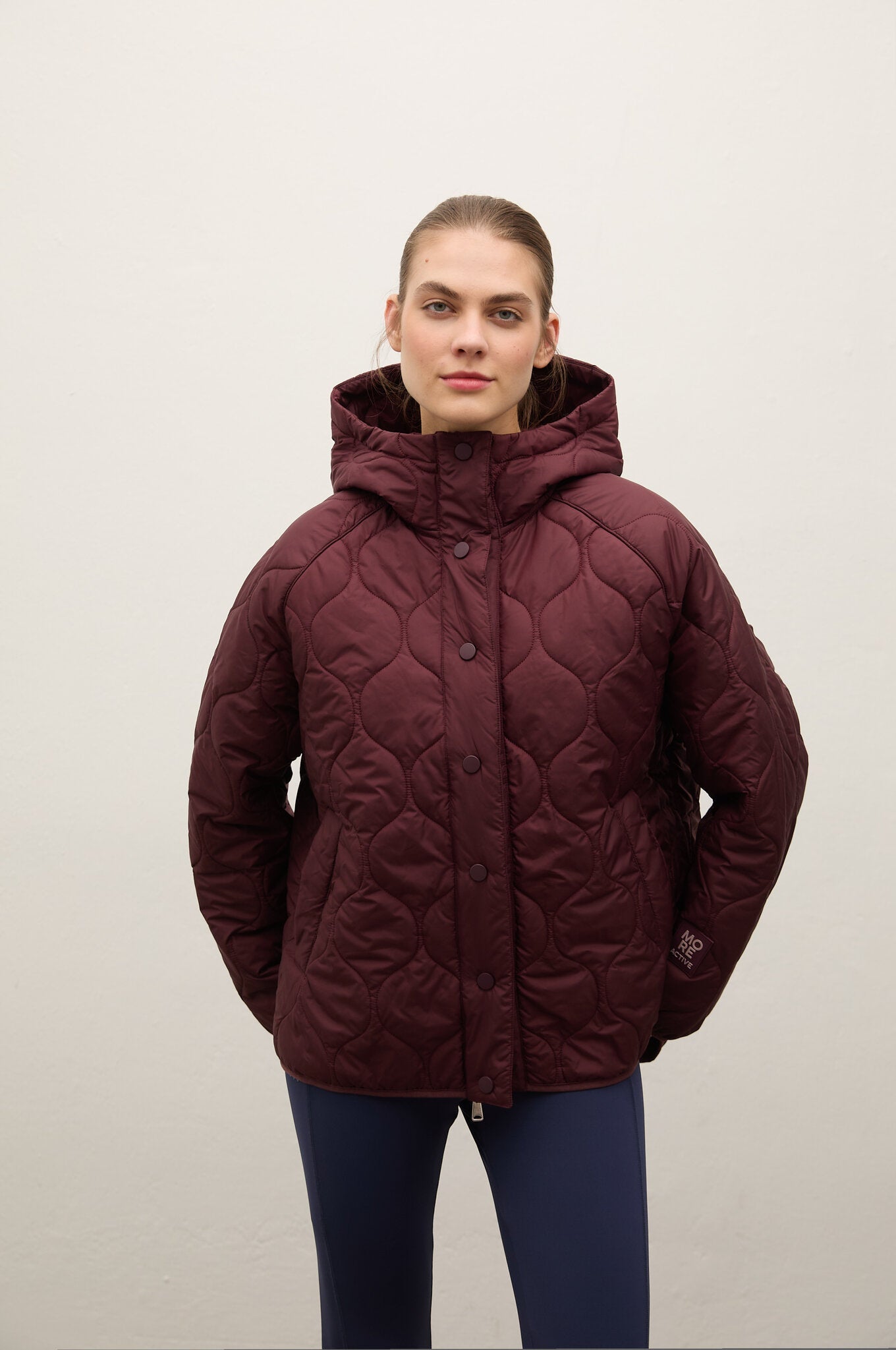 A woman modeling a quilted hooded puffer jacket in a deep burgundy color, paired with navy blue leggings, against a plain off-white background.