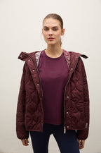 A woman modeling a dark burgundy quilted puffer jacket over a matching burgundy t-shirt and navy pants, standing against a plain off-white background.