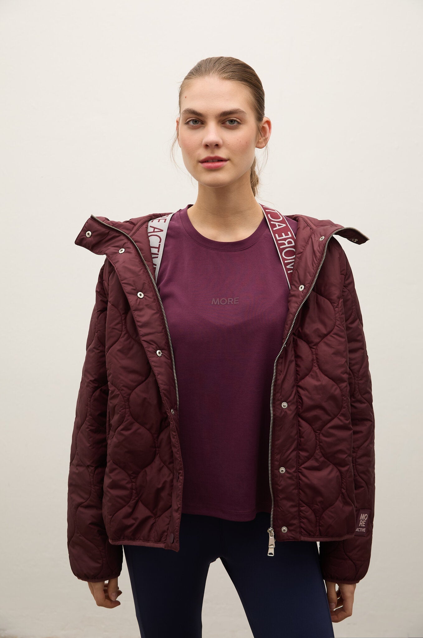 A woman modeling a dark burgundy quilted puffer jacket over a matching burgundy t-shirt and navy pants, standing against a plain off-white background.