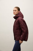 A woman wearing a quilted puffer jacket in deep burgundy with a high collar, standing in profile against a plain off-white wall.