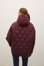 A woman viewed from behind wearing a quilted maroon hooded puffer jacket and dark blue leggings against a plain off-white wall.