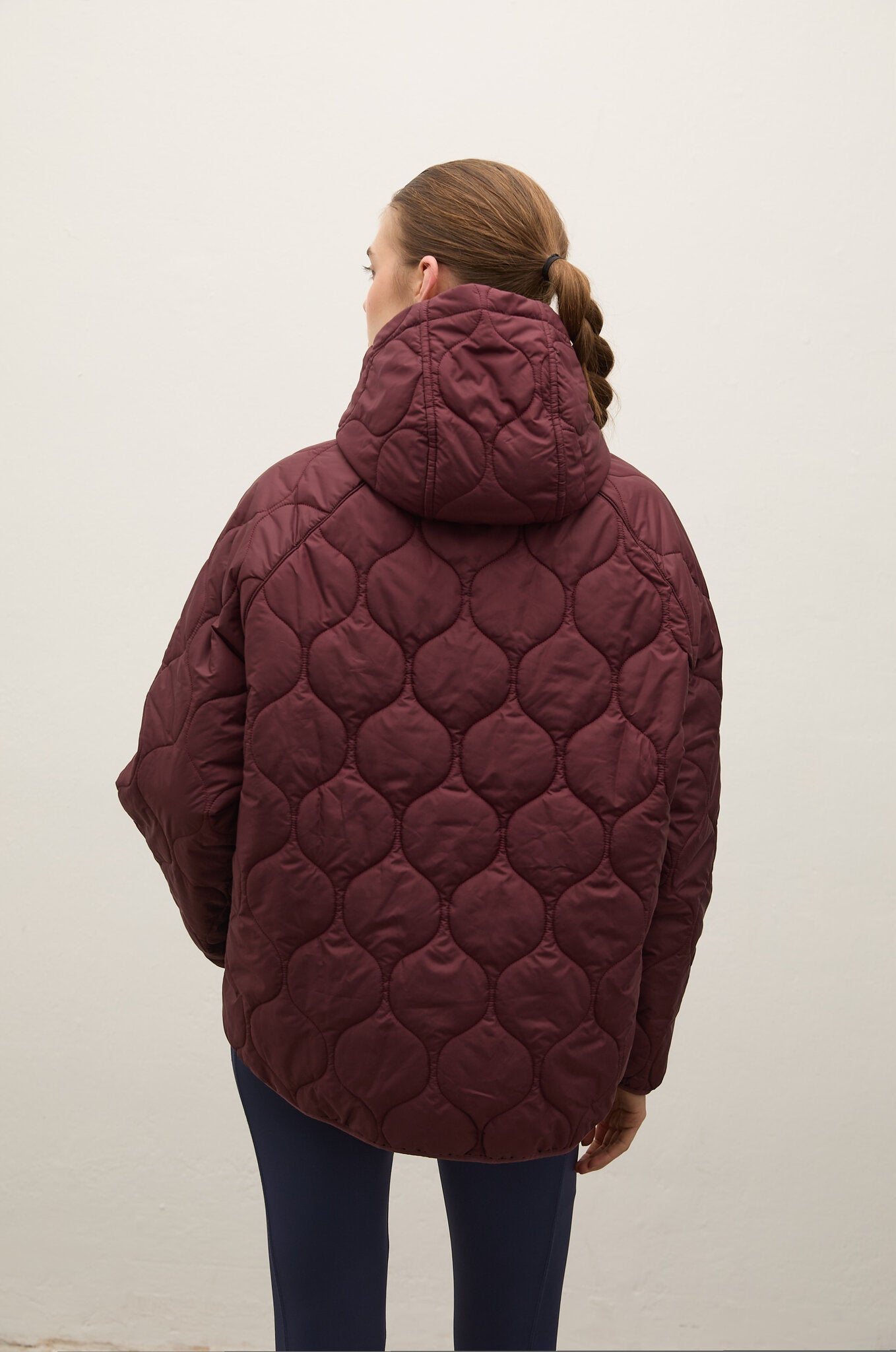 A woman viewed from behind wearing a quilted maroon hooded puffer jacket and dark blue leggings against a plain off-white wall.