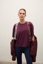 A woman modeling a short-sleeve athletic t-shirt in deep burgundy, paired with dark blue leggings and carrying a matching burgundy puffer jacket over her shoulders. The t-shirt features a small white 'MORI' logo on the chest and white text on the shoulder straps reading 'MORE ACTIVE'.