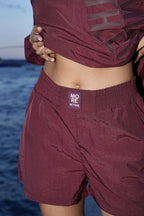 A close-up of a person wearing burgundy athletic shorts with an elastic waistband and a visible 'MORE ACTIVE' label, paired with a matching cropped top, against an outdoor water background.
