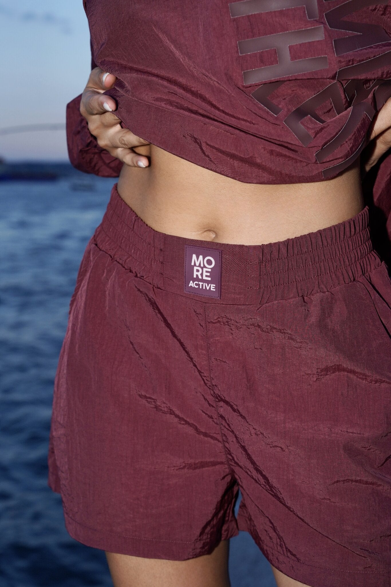 A close-up of a person wearing burgundy athletic shorts with an elastic waistband and a visible 'MORE ACTIVE' label, paired with a matching cropped top, against an outdoor water background.