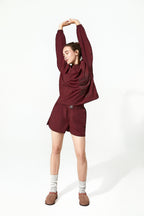 A woman wearing a matching burgundy lounge set consisting of a long-sleeve top and shorts, styled with white socks and brown slippers against a plain white studio background.