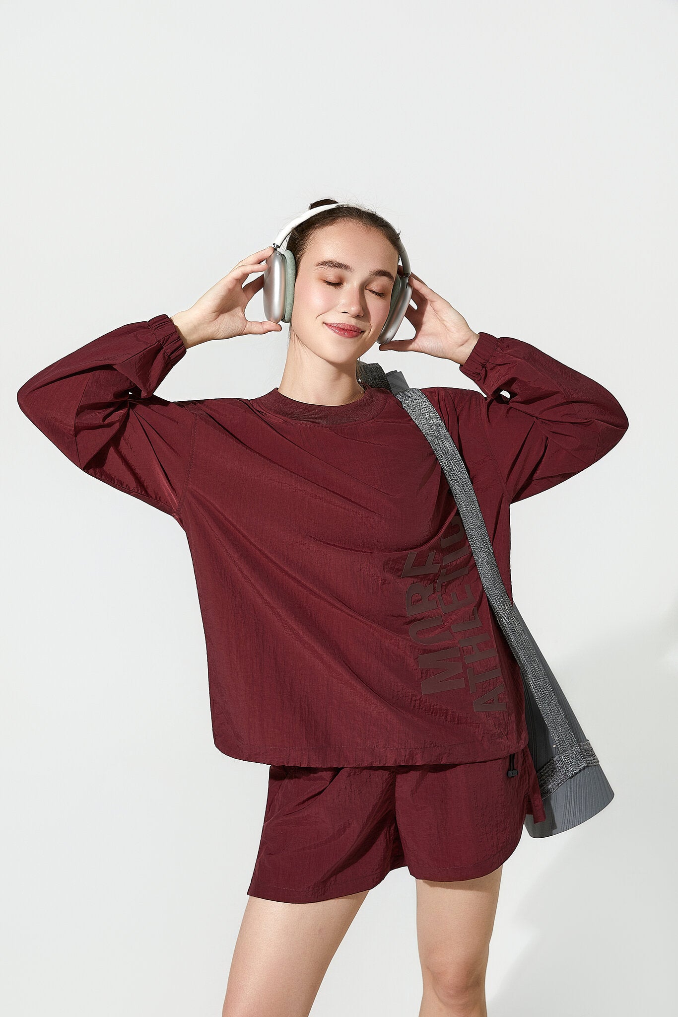 A woman wearing a matching burgundy long-sleeve top and shorts set, with a gray shoulder bag and silver headphones, posing against a light gray studio background.