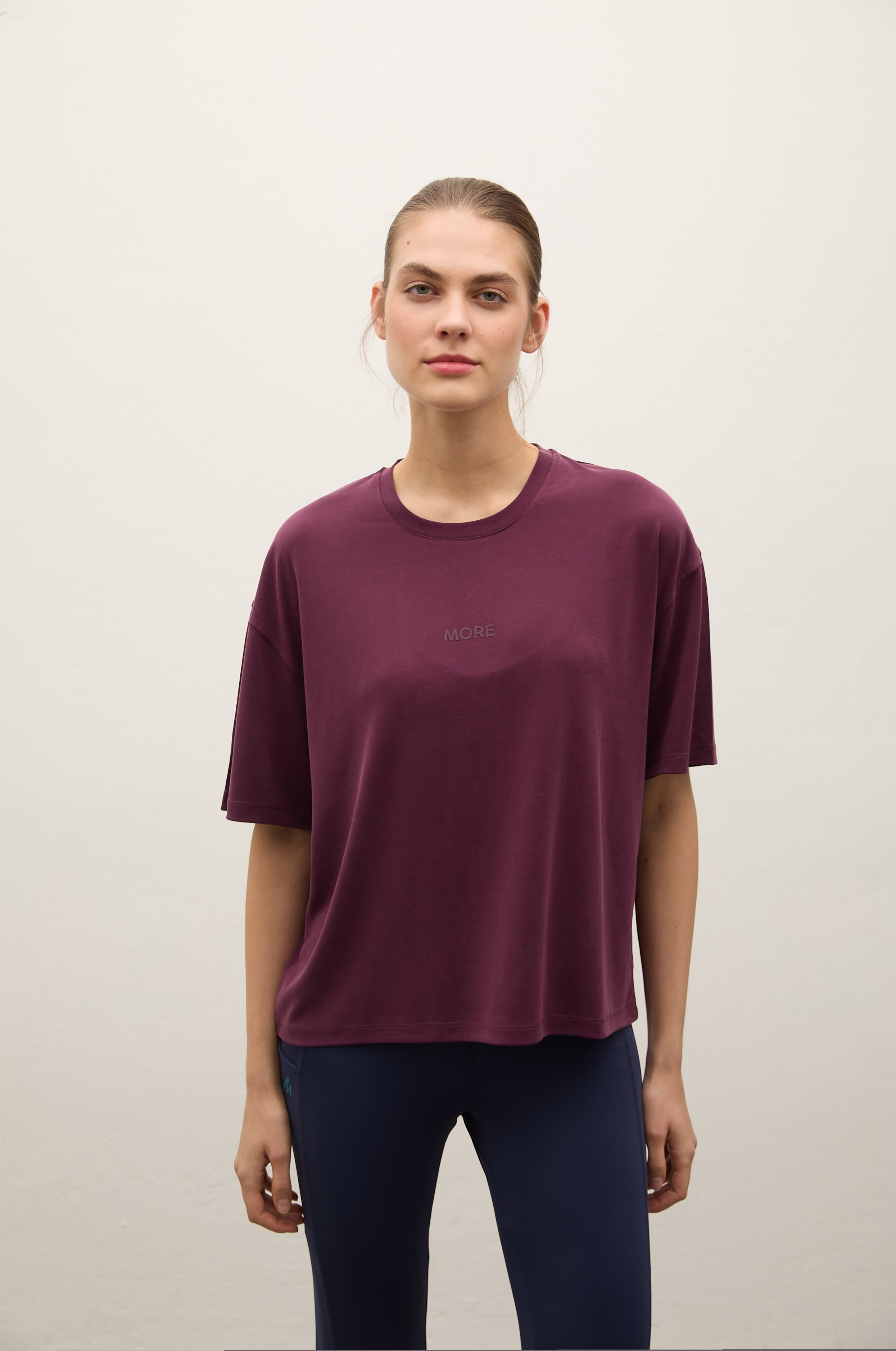 A woman modeling an oversized short-sleeve crewneck t-shirt in deep burgundy, paired with dark navy leggings, against a plain off-white studio background.