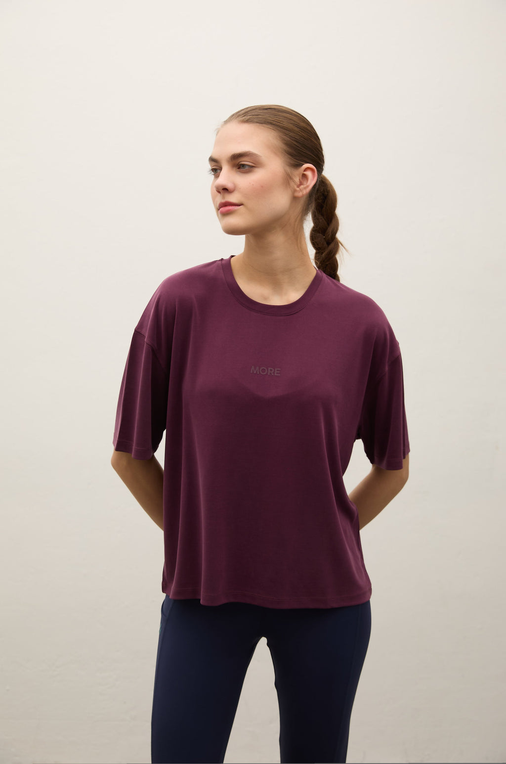 A woman wearing a short-sleeve crewneck t-shirt in deep plum purple, paired with dark navy leggings, standing against a plain off-white wall.