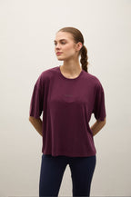 A woman wearing a short-sleeve crewneck t-shirt in deep plum purple, paired with dark navy leggings, standing against a plain off-white wall.