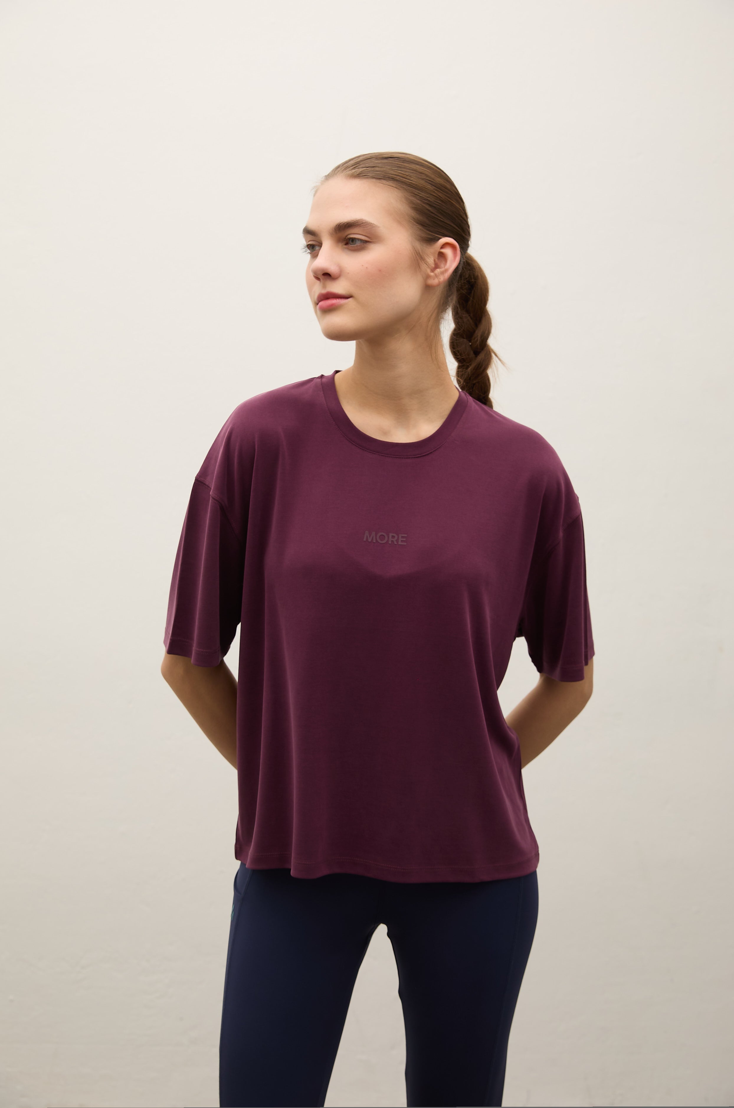 A woman wearing a short-sleeve crewneck t-shirt in deep plum purple, paired with dark navy leggings, standing against a plain off-white wall.