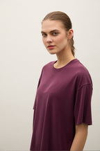 A woman with her hair pulled back wears an oversized short-sleeve crewneck t-shirt in a muted plum purple color. The shirt has a small tonal embroidered logo on the left chest.