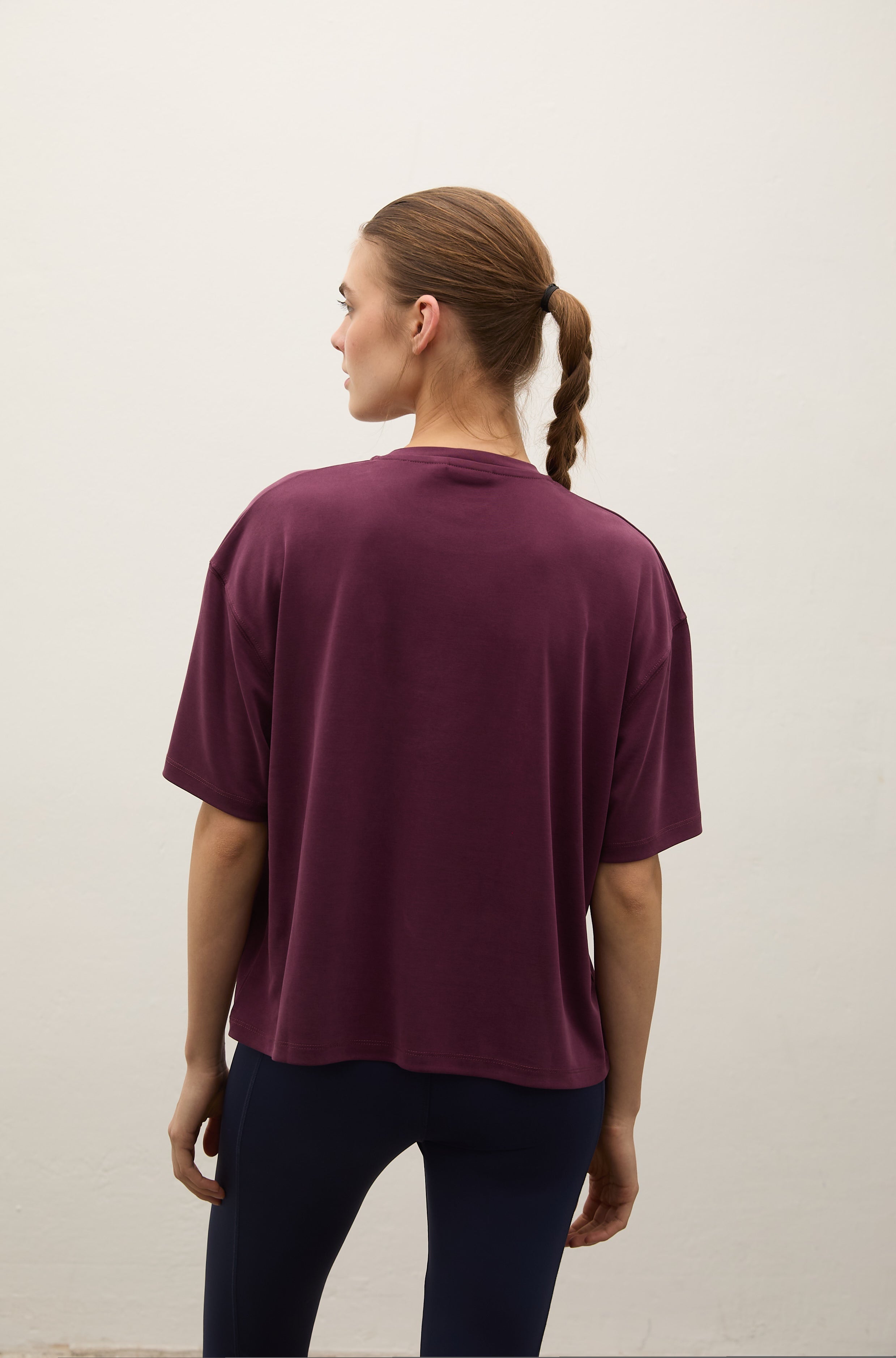 A woman viewed from behind wearing a loose, short-sleeve crewneck t-shirt in deep plum purple and dark navy leggings, standing against a plain off-white wall.