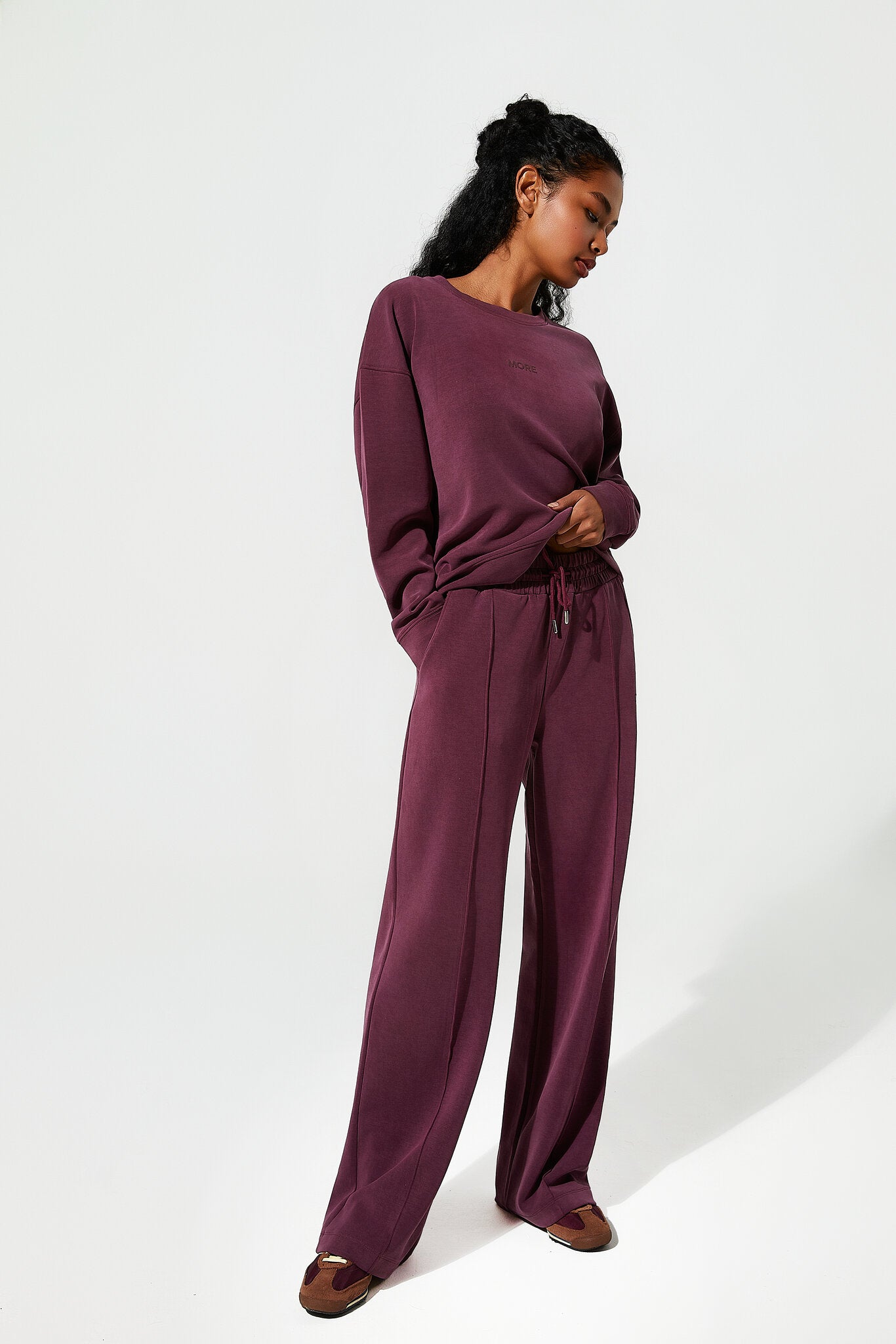 A woman modeling a matching two-piece lounge set in a burgundy color, consisting of a long-sleeve top and wide-leg pants, against a plain white studio background.