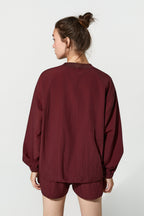 A woman shown from the back wearing a loose-fitting burgundy long-sleeve top and matching shorts against a plain light gray studio background.