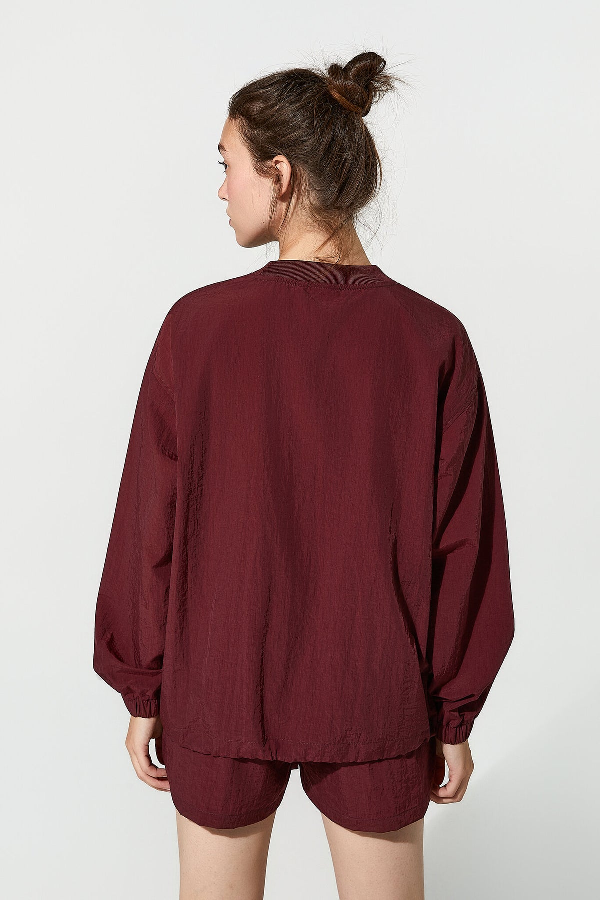 A woman shown from the back wearing a loose-fitting burgundy long-sleeve top and matching shorts against a plain light gray studio background.