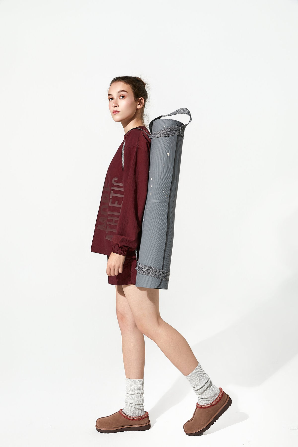 A woman in a side profile wearing a burgundy athletic sweatshirt, shorts, and brown slip-on shoes, carrying a rolled-up gray yoga mat on her back against a white studio background.