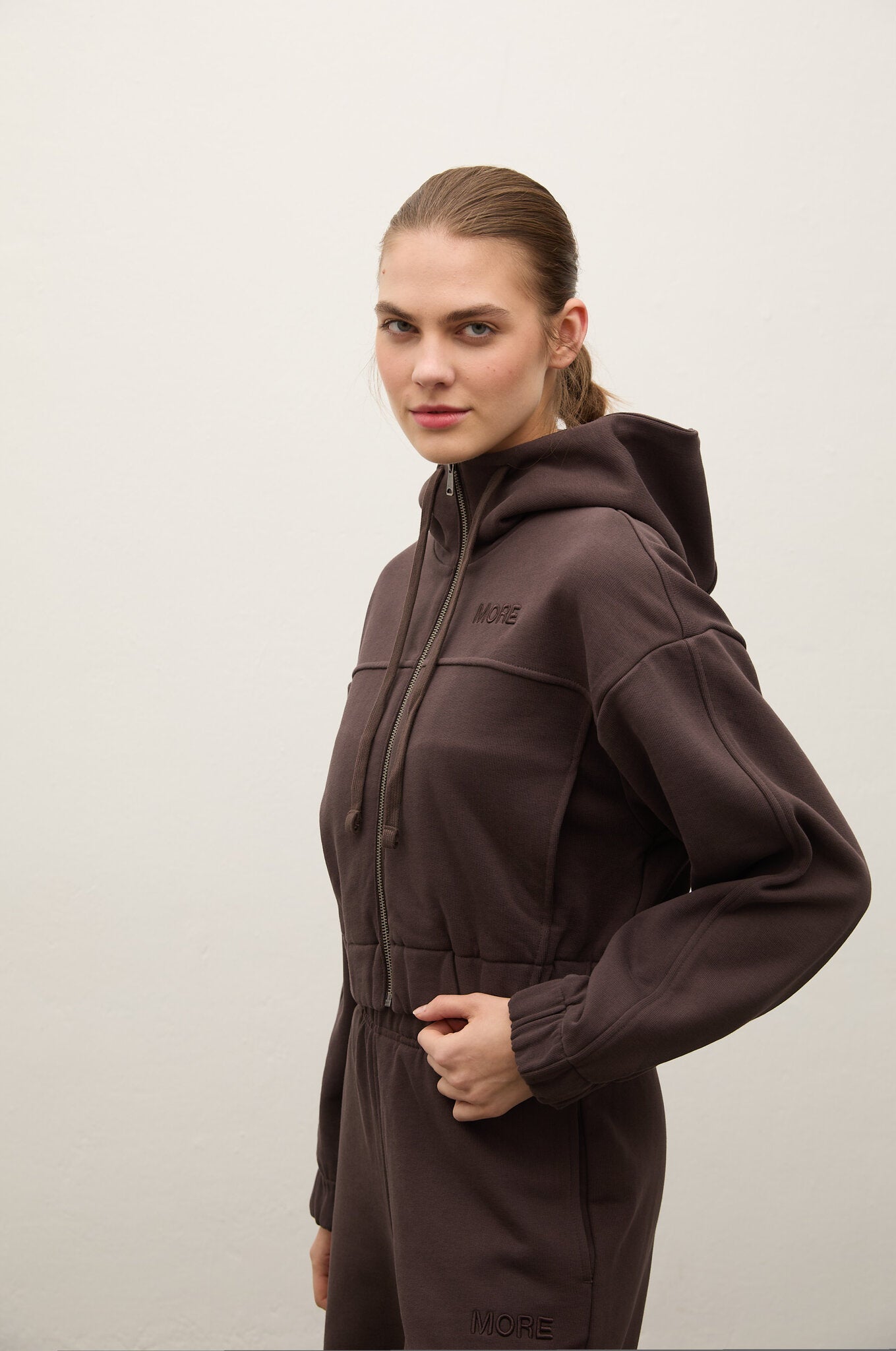 A woman modeling a dark brown hooded zip-up sweatshirt and matching sweatpants against a plain light gray background.