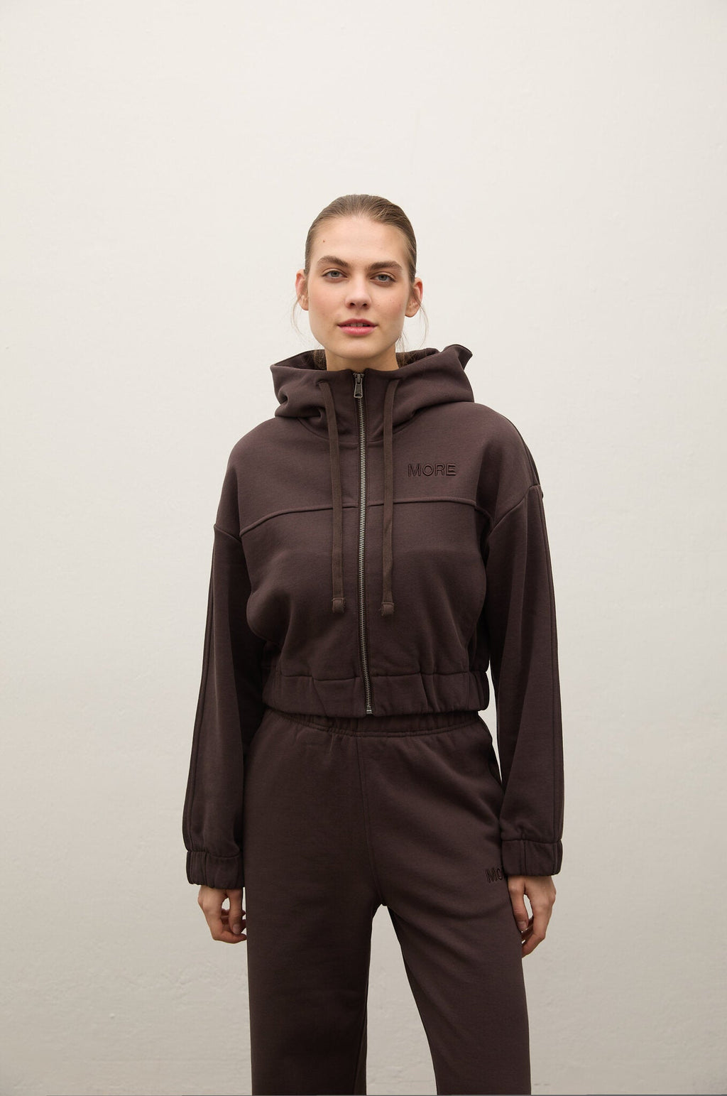A model wearing a dark brown cropped zip-up hoodie with a drawstring hood and matching dark brown sweatpants, standing against a plain light gray wall.