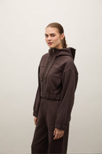 A woman modeling a dark brown hooded jumpsuit with a front zipper and matching pants, standing against a plain light gray studio background.