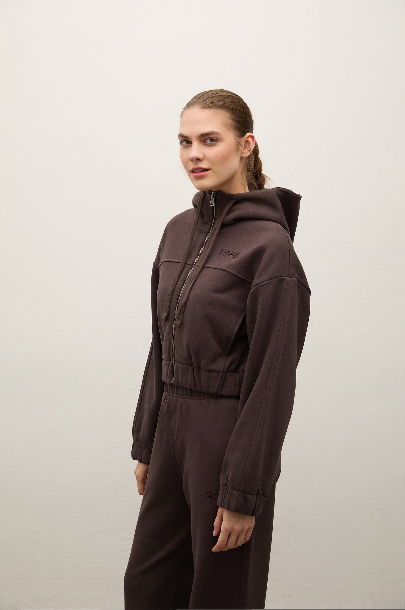 A woman modeling a dark brown hooded jumpsuit with a front zipper and matching pants, standing against a plain light gray studio background.