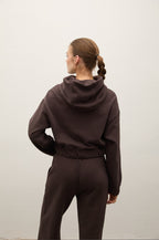 A woman viewed from behind wearing a dark brown hooded sweatshirt and matching jogger pants against a plain light gray wall.