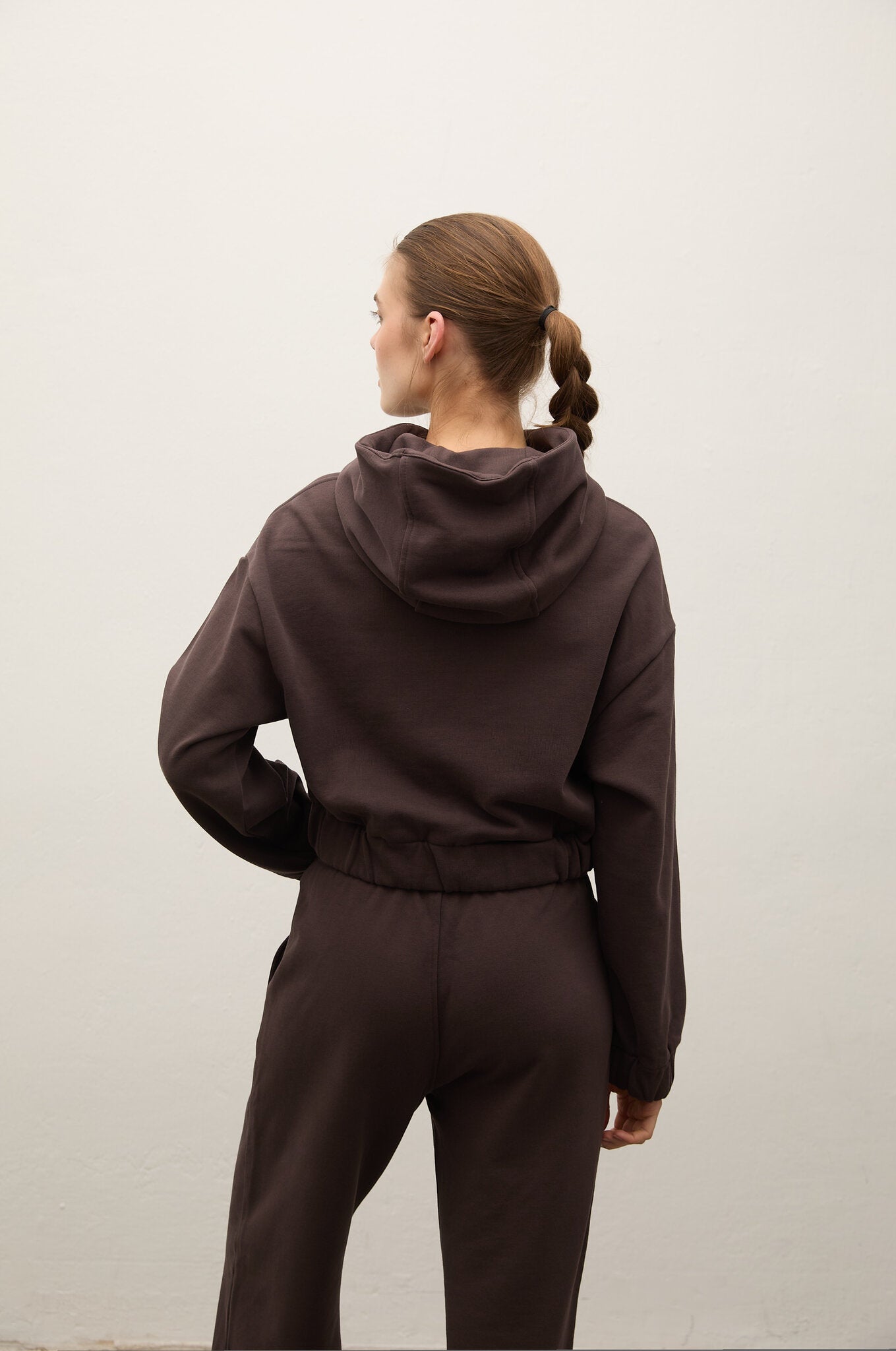 A woman viewed from behind wearing a dark brown hooded sweatshirt and matching jogger pants against a plain light gray wall.