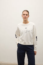 A woman modeling a cream-colored crewneck sweatshirt with graphic prints on the chest and sleeves, paired with dark navy pants, standing against a plain off-white wall.