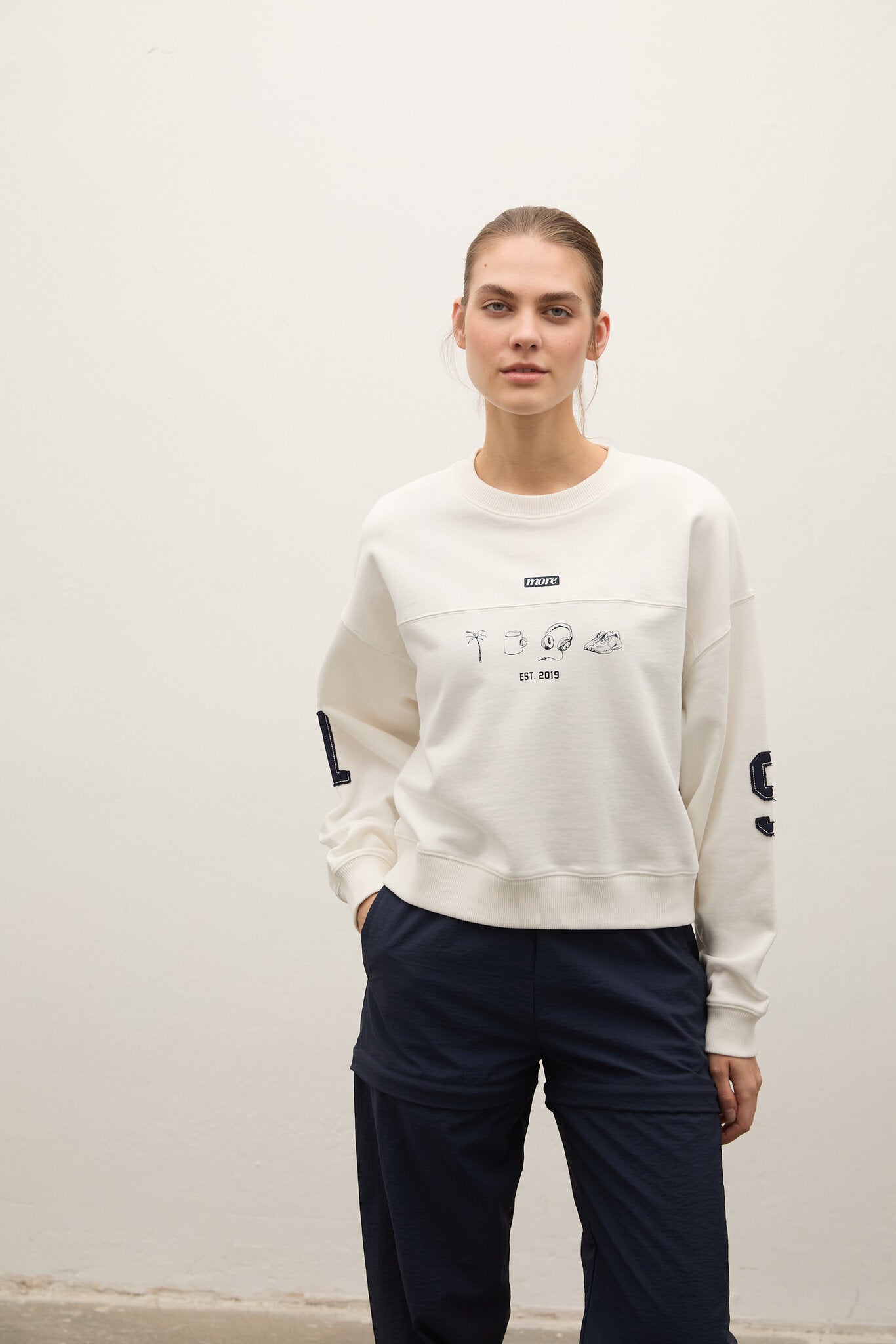 A woman modeling a cream-colored crewneck sweatshirt with graphic prints on the chest and sleeves, paired with dark navy pants, standing against a plain off-white wall.