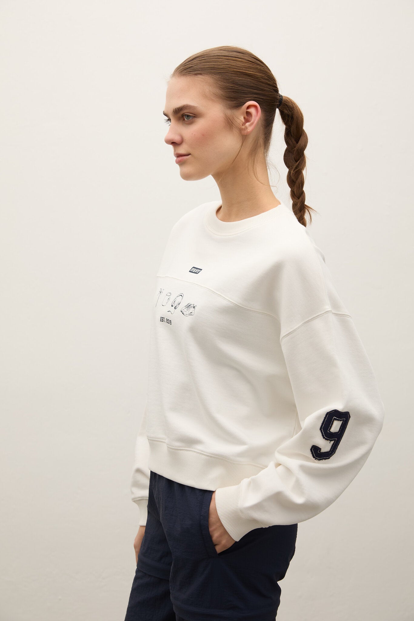 Woman wearing a white sweatshirt with a number 9 on the sleeve against a plain background