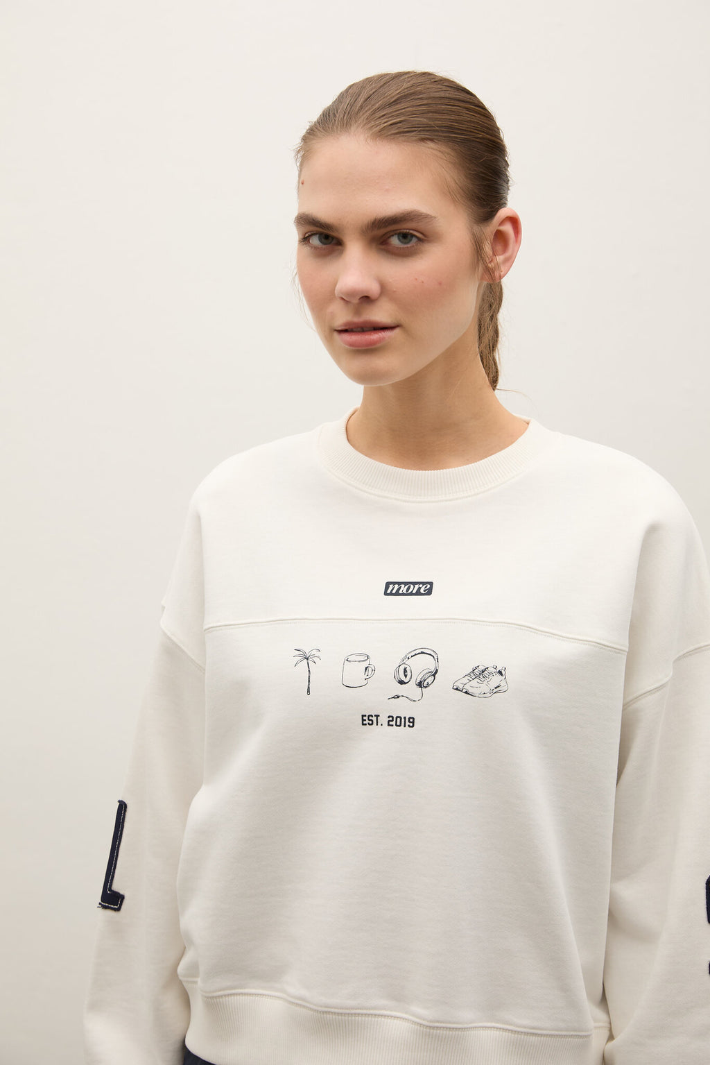 A woman wearing a cream-colored crewneck sweatshirt with black graphic illustrations and text on the front, photographed against a plain light background.