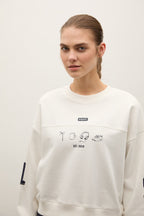 A woman wearing a cream-colored crewneck sweatshirt with black graphic illustrations and text on the front, photographed against a plain light background.