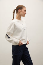 A woman wearing a white cropped sweatshirt with a graphic print on the chest and a navy sleeve patch, paired with dark navy pants, standing against a plain off-white studio background.