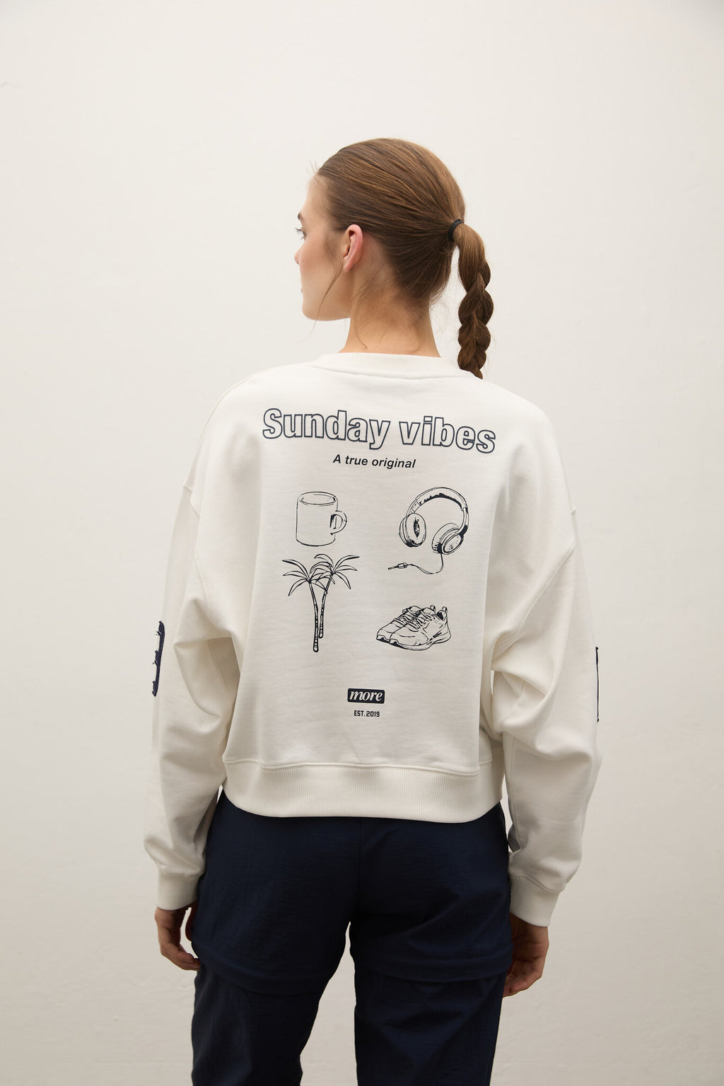 Person wearing a light gray sweatshirt with 'Sunday vibes' text and graphics on the back, standing against a plain background.
