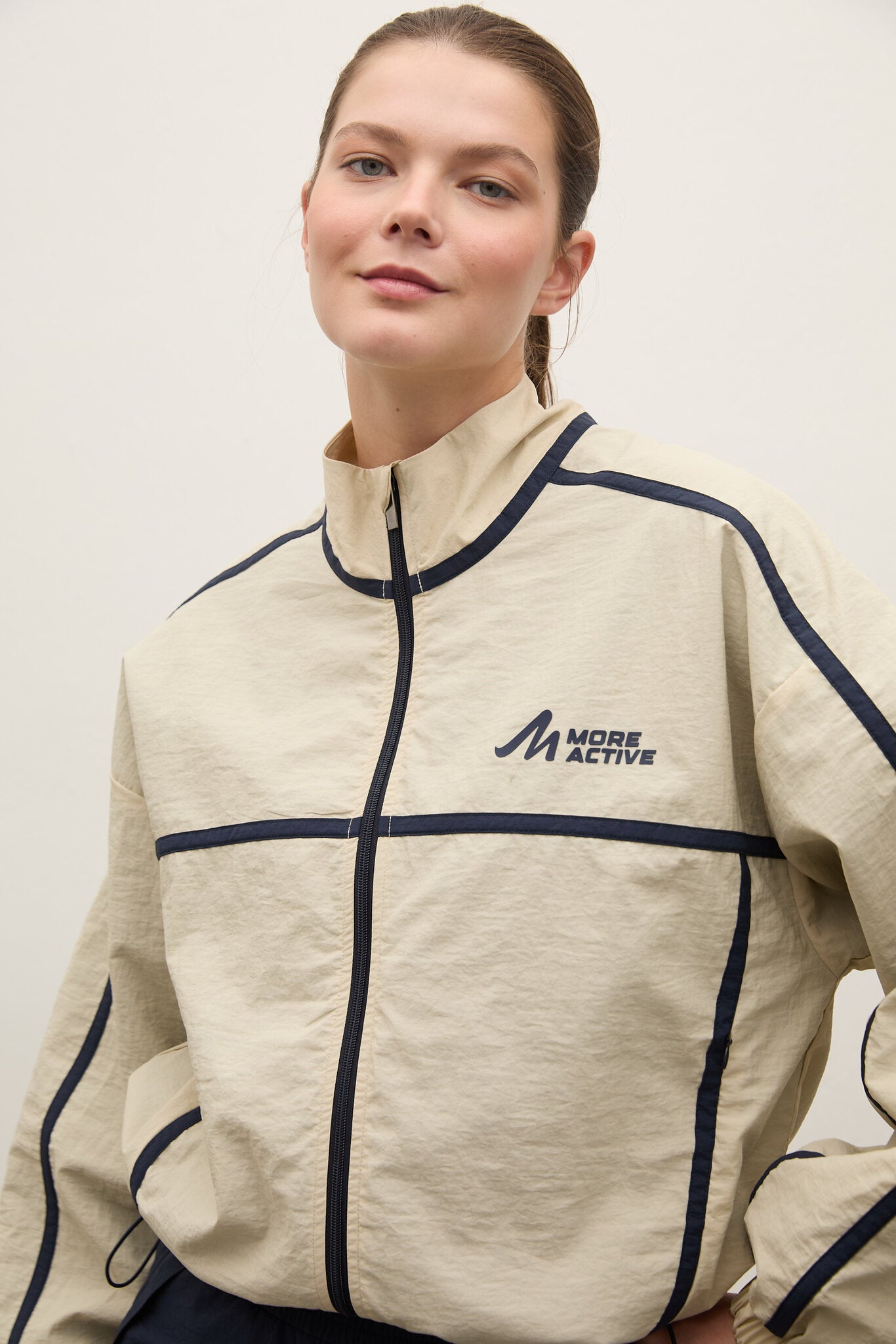 A woman wearing a beige zip-up athletic jacket with navy blue piping and a 'MORE ACTIVE' logo on the chest, photographed against a plain light background.