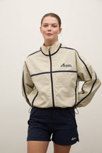 A woman modeling a beige athletic zip-up jacket with dark navy contrast piping and a dark navy logo on the chest, paired with dark navy athletic shorts against a plain light background.