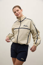 A woman wearing a beige athletic zip-up jacket with dark navy piping and a matching dark navy athletic shorts, posed against a plain off-white studio background.