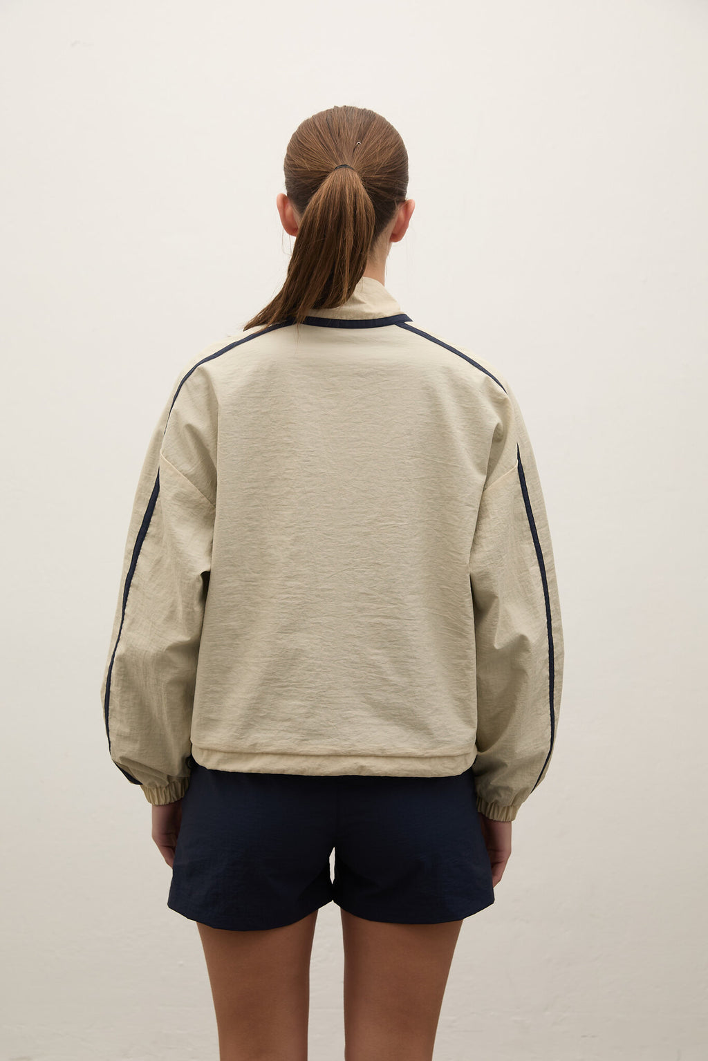 A person viewed from the back wearing a light beige cropped jacket with dark navy piping along the shoulders and sleeves, paired with dark navy shorts, against a plain off-white wall.