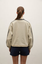 A person viewed from the back wearing a light beige cropped jacket with dark navy piping along the shoulders and sleeves, paired with dark navy shorts, against a plain off-white wall.