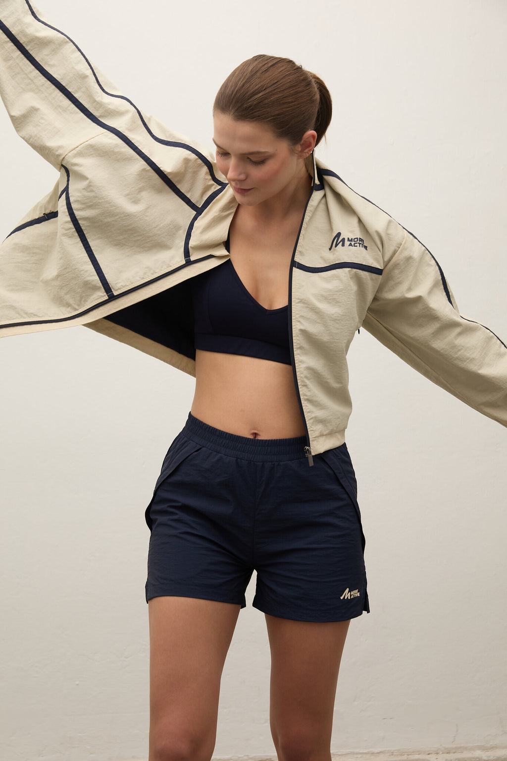 Woman wearing a beige jacket, black sports bra, and navy shorts on a plain background