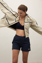 Woman wearing a beige jacket, black sports bra, and navy shorts on a plain background