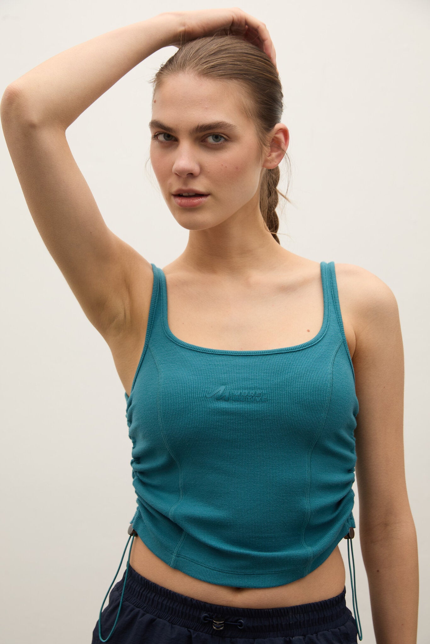 A woman modeling a teal athletic crop top with side drawstrings, paired with dark navy athletic bottoms, against a plain light gray background.