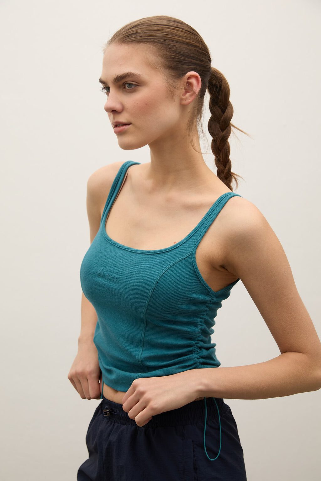 A woman wearing a teal ruched scoop-neck tank top and dark navy drawstring pants, photographed against a plain light gray studio background.