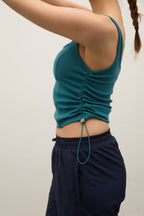 A side-profile view of a person wearing a teal ruched tank top and dark navy pants against a plain light background.
