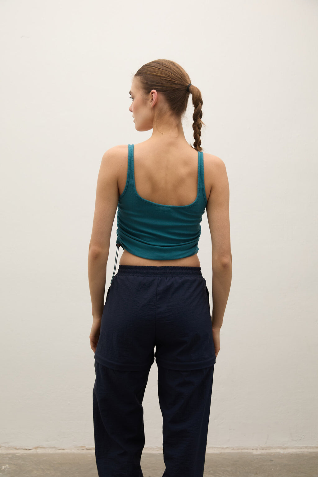 Person wearing a green tank top and navy pants against a white wall