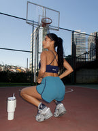 A woman crouches on an outdoor basketball court wearing a navy blue sports bra and teal athletic shorts, with a white tumbler nearby and a basketball hoop in the background.