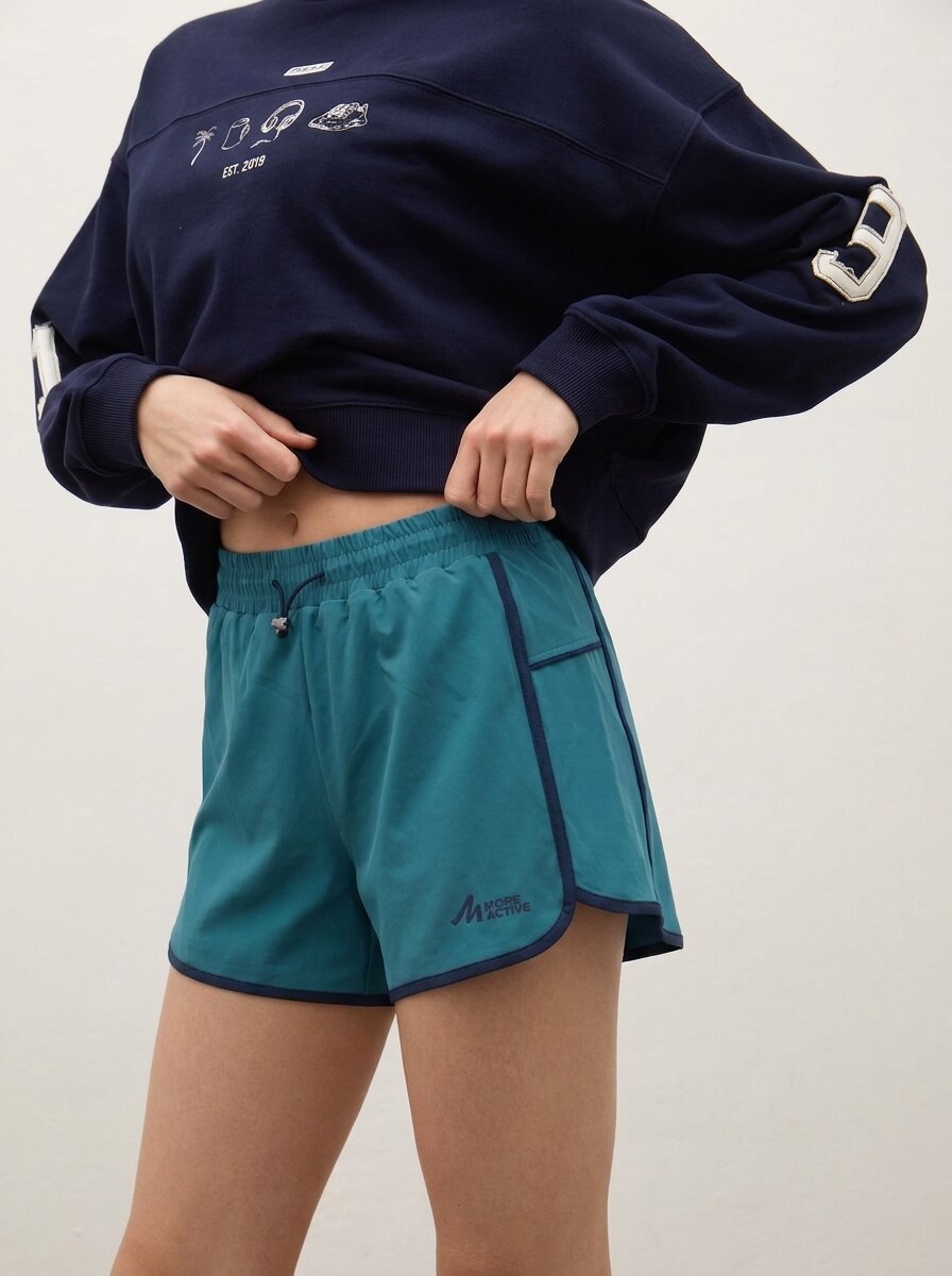 A model wearing teal athletic shorts with dark navy trim and a matching navy sweatshirt, cropped from the chest down against a plain light background.
